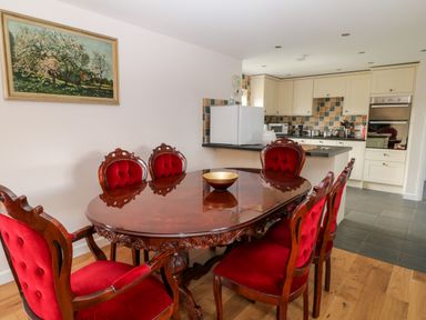 A dining room with a wooden table and red upholstered chairs next to a kitchen with cream cabinets and tiled floor at Honeysuckle Cottage Ashwater near Holsworthy