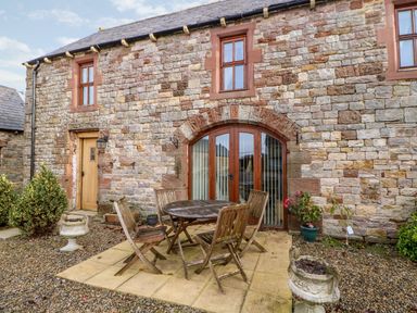 An outdoor patio with a round wooden table and four wooden chairs in front of a stone building with arched glass doors and two windows at The Hayloft in Milton near Brampton