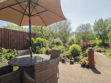 An outdoor patio with a round glass table, wicker chairs, a large umbrella, and surrounding garden plants at Kingfisher Wattisfield near Rickinghall and Diss