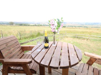 A wooden table with a bottle of prosecco two glasses and a vase of flowers on a wooden deck with a field view at The Shooting Lodge in Longnor