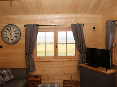 A wooden room with a window in the center brown curtains a wall clock a TV on a cabinet and a sofa with a side table at The Shooting Lodge in Longnor
