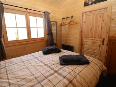 A bedroom with a bed covered in plaid bedding towels on bed wooden walls window with curtains coat hangers on wall and wooden door at The Shooting Lodge in Longnor