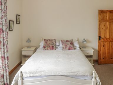 A bedroom with a white bed frame white bedding floral pillows two bedside tables with lamps floral curtains wooden door at Seaspray in Tully County Galway