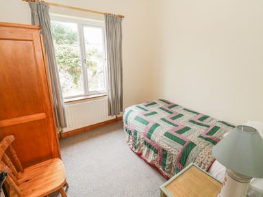 A bedroom with a single bed covered with a quilt next to a nightstand with a lamp a wooden chair and wardrobe near a window with curtains at Seaspray in Tully County Galway