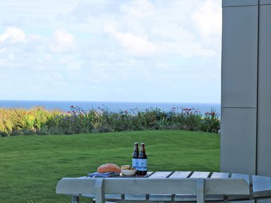 A wooden table with two bottles, bread, and a bowl with sea and garden in the background at Saltwater, One Lusty Glaze in Newquay
