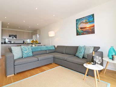 A living room with a gray sectional sofa and blue pillows next to a kitchen with cabinets at Saltwater One Lusty Glaze in Newquay