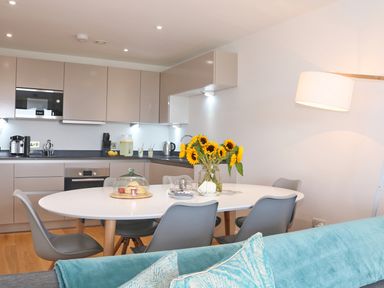 A kitchen and dining area with a white table and gray chairs a vase of sunflowers and snacks under a glass cover at Saltwater One Lusty Glaze in Newquay