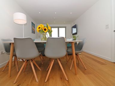 A dining area with a white table and six gray chairs with wooden legs a vase of sunflowers on the table a floor lamp in the corner a wall-mounted television and white walls at Saltwater One Lusty Glaze in Newquay