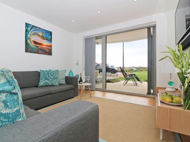 A living room with grey sofas and blue cushions next to sliding glass doors opening to a patio with chairs at Saltwater One Lusty Glaze in Newquay