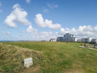 A large grassy field with a stone marker and modern buildings in the background at Saltwater One Lusty Glaze in Newquay