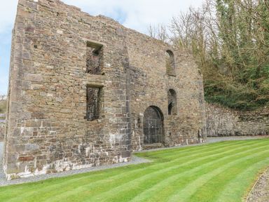 A stone ruin with arched windows and a barred entrance next to a lawn and trees at Heddwch Wisemans Bridge near Saundersfoot
