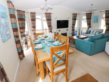 A dining area with a wooden table and six chairs set for a meal next to a living room with sofas and a wall mounted tv at Heddwch Wisemans Bridge near Saundersfoot
