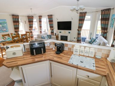 A kitchen counter with appliances and containers overlooking a dining and living area with curtains and a wall mounted television at Heddwch Wisemans Bridge near Saundersfoot