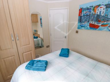 A bedroom with a bed covered in a light checkered duvet and blue towels on top a wardrobe a door a wall mirror and a painting of boats on the wall at Heddwch Wisemans Bridge near Saundersfoot