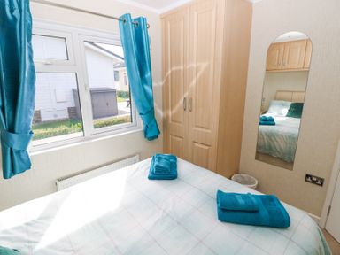 A bedroom with a bed holding folded towels next to a wooden wardrobe and a window with blue curtains at Heddwch Wisemans Bridge near Saundersfoot