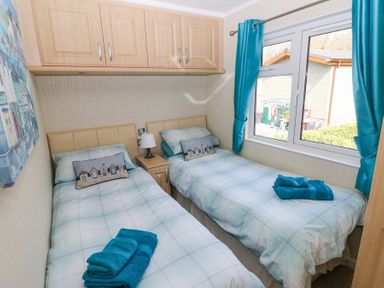 A bedroom with two single beds, a nightstand with a lamp, overhead cabinets, and a window with blue curtains at Heddwch in Wisemans Bridge near Saundersfoot
