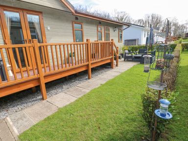 A wooden deck attached to a house with a pathway and grassy lawn with bird feeders and outdoor seating area at Heddwch Wisemans Bridge near Saundersfoot