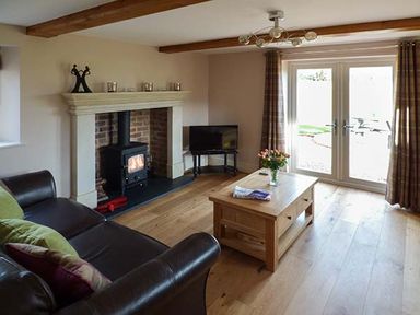 A living room with a wood burning stove a brown leather sofa a wooden coffee table with flowers and magazines and glass doors with curtains at Rose Cottage in Winforton near Hay-on-Wye