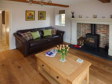 A living room with a brown leather sofa green and plaid cushions a wooden coffee table with flowers books and a fireplace with wood logs at Rose Cottage in Winforton near Hay-on-Wye