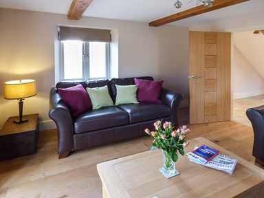 A living room with a brown leather sofa with cushions a wooden coffee table with flowers and papers a lamp on a side table and an open door at Rose Cottage in Winforton near Hay-on-Wye