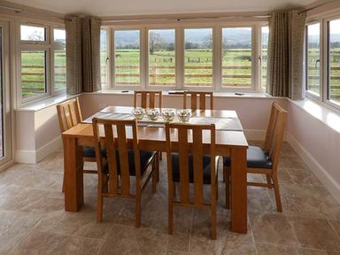 A dining area with a wooden table and six chairs surrounded by windows at Rose Cottage in Winforton near Hay-on-Wye