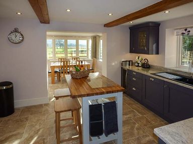 A kitchen with wooden island and stools next to dark cabinets and a dining area with wooden table and chairs at Rose Cottage in Winforton near Hay-on-Wye