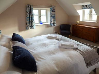 A bedroom with a bed with pillows and towels near two windows with curtains a dark chair and a wooden dresser at Rose Cottage in Winforton near Hay-on-Wye