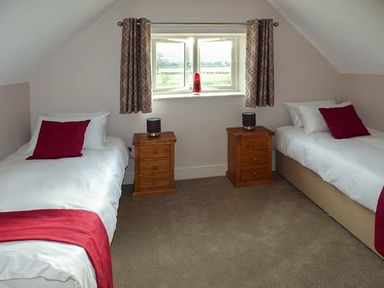 A bedroom with two single beds each with white bedding and red pillows and throws two wooden bedside tables with lamps and a window with patterned curtains