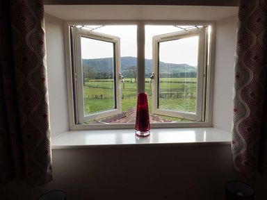 A window with two open panes and a red vase on the sill with green fields and hills outside at Rose Cottage in Winforton near Hay-on-Wye