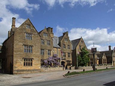 A large stone building with multiple gables windows flags and a flowering bush along a road at Kitts Cottage in Little Malvern near Malvern