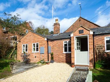 A brick cottage with a half open door a small patio with a table and chairs and a gravel driveway at Kitts Cottage in Little Malvern near Malvern