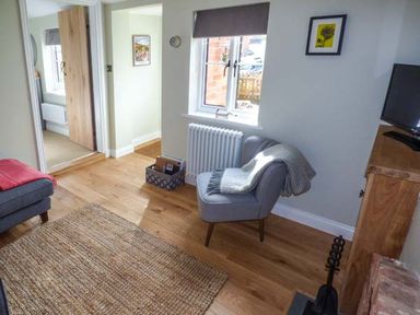 A sitting room with a gray chair and blanket a wooden floor a woven rug and a television at Kitts Cottage in Little Malvern near Malvern