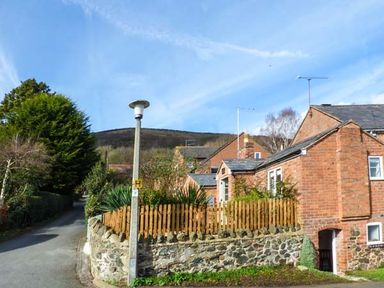 A brick house with a stone wall and wooden fence beside a street with trees at Kitts Cottage in Little Malvern near Malvern
