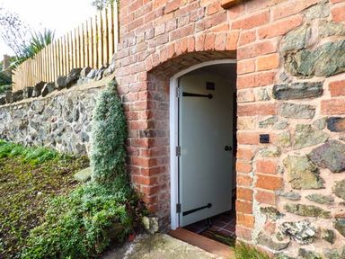 A half-open door set in a brick and stone wall with plants growing near the entrance at Kitts Cottage in Little Malvern near Malvern