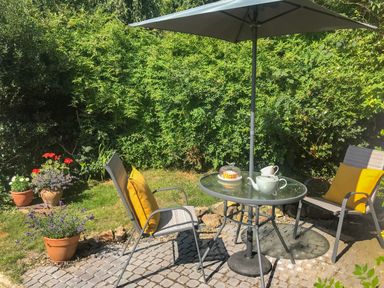 A patio area with two chairs with yellow cushions a glass table with a teapot and cake and an umbrella surrounded by greenery at Kitts Cottage in Little Malvern near Malvern