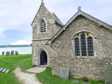 A stone church building with arched windows and entrance near gravestones and water at Kilcarreg Rock near Wadebridge