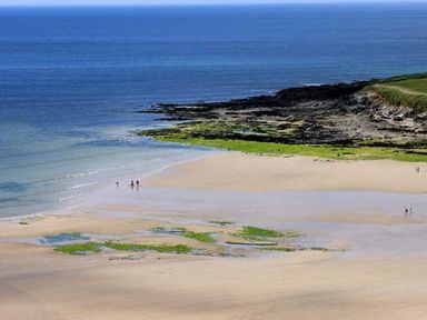 A sandy beach with some people walking near the water and rocky areas with green seaweed at Kilcarreg Rock near Wadebridge