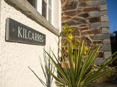 A black sign with Kilcarreg on a white wall next to a window and a green plant outside the building at Kilcarreg Rock near Wadebridge