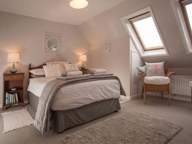 A bedroom with a double bed and bedside tables with lamps and a wicker chair with cushions near skylight windows at Kilcarreg Rock near Wadebridge