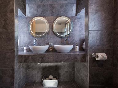 A bathroom with two round mirrors with lights above two white sinks and a toilet paper holder on the wall at Kilcarreg Rock near Wadebridge
