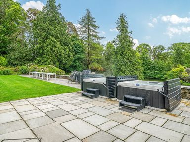 An outdoor area with two hot tubs on a stone patio surrounded by grass and trees at The Oaks in Bowness & Windermere