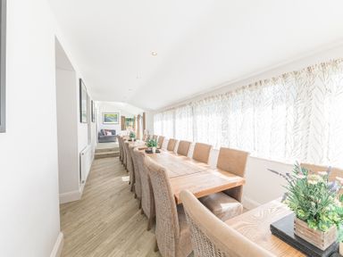 A long dining room with a wooden table and chairs next to windows with sheer curtains at The Oaks in Bowness & Windermere