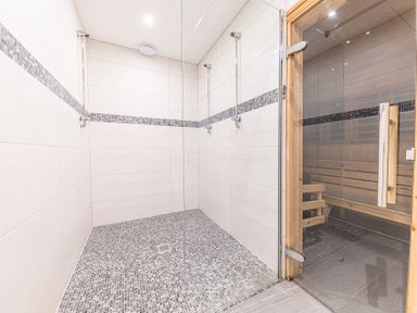 A tiled shower area with two shower heads and a glass door leading to a wooden sauna at The Oaks in Bowness & Windermere