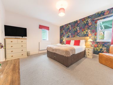 A bedroom with a double bed floral wallpaper two windows a chest of drawers and a wall-mounted television at The Oaks in Bowness & Windermere