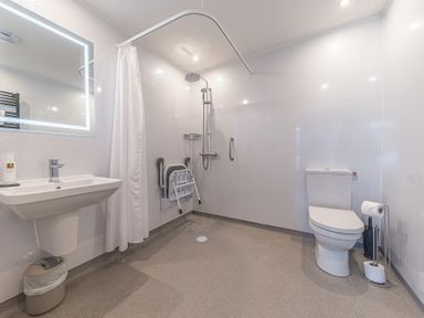 A bathroom with a sink mirror and trash bin a shower area with a curtain foldable seat and grab bar and a toilet with toilet paper holders at The Oaks in Bowness & Windermere