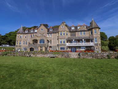 A large stone building with multiple windows and pitched roofs with turrets with a grassy lawn in front at Menai View near Menai Bridge