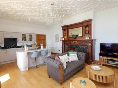 An open living room and kitchen with a sofa, wooden coffee tables, a television, a kitchen island with stools, and a decorative fireplace at Menai View near Menai Bridge