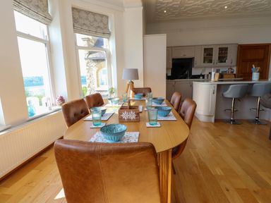 A dining area with a wooden table set with blue bowls and glasses and brown chairs next to large windows and an open kitchen at Menai View near Menai Bridge