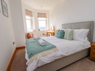 A bedroom with a bed covered with white sheets green pillows and a green throw near windows and wooden furniture at Menai View near Menai Bridge