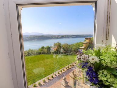 A window view overlooking a green lawn with a flower arrangement on the right and a body of water beyond with hills in the background at Menai View near Menai Bridge